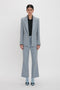 Person standing against a plain white background, impeccably tailored in a light gray Exclusive Sleeve Detail Patch Pocket Jacket In Marina by Victoria Beckham with contemporary detailing, featuring a black shirt and light blue socks paired with black and blue shoes.