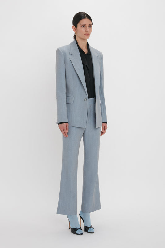 A person stands against a plain white background, wearing an impeccably tailored Exclusive Sleeve Detail Patch Pocket Jacket In Marina by Victoria Beckham with contemporary detailing, paired with a black blouse and matching blue heels.