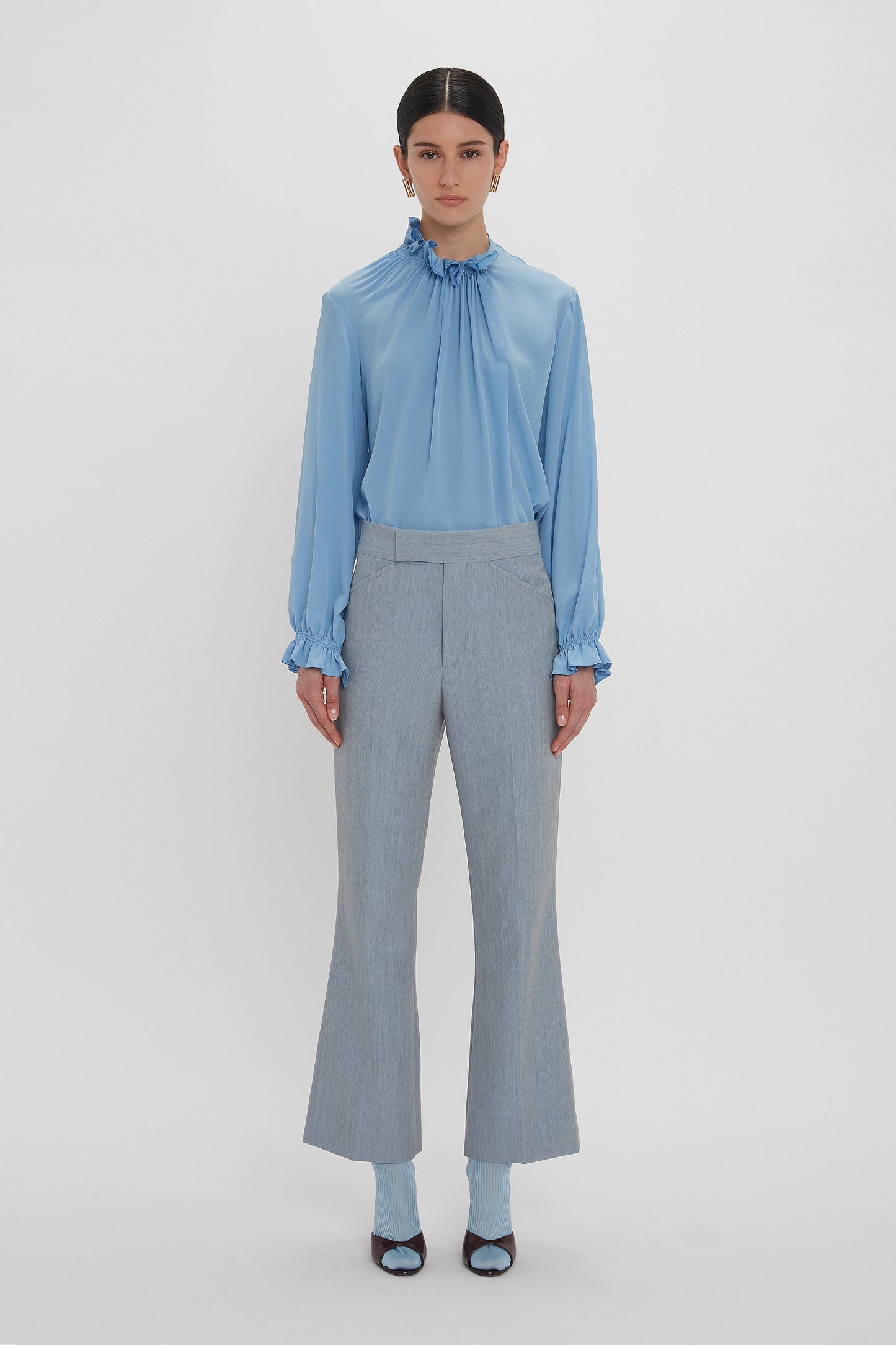 A person stands against a plain white background, wearing an Exclusive Ruffle Neck Blouse In Cornflower Blue by Victoria Beckham made of crepe de chine fabric with matching high-waisted, light gray pants featuring flared legs and blue socks paired with black open-toe heels.