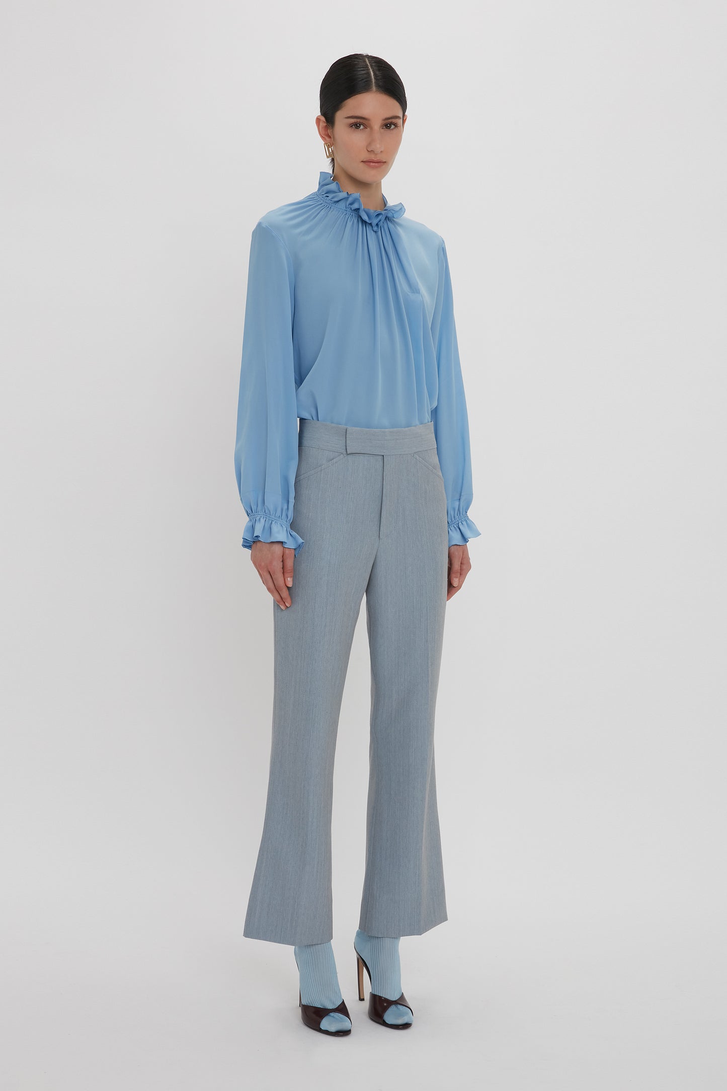 A person stands against a plain background, wearing the Victoria Beckham Exclusive Ruffle Neck Blouse In Cornflower Blue, paired with high-waisted, light gray pants and matching cornflower blue heels.