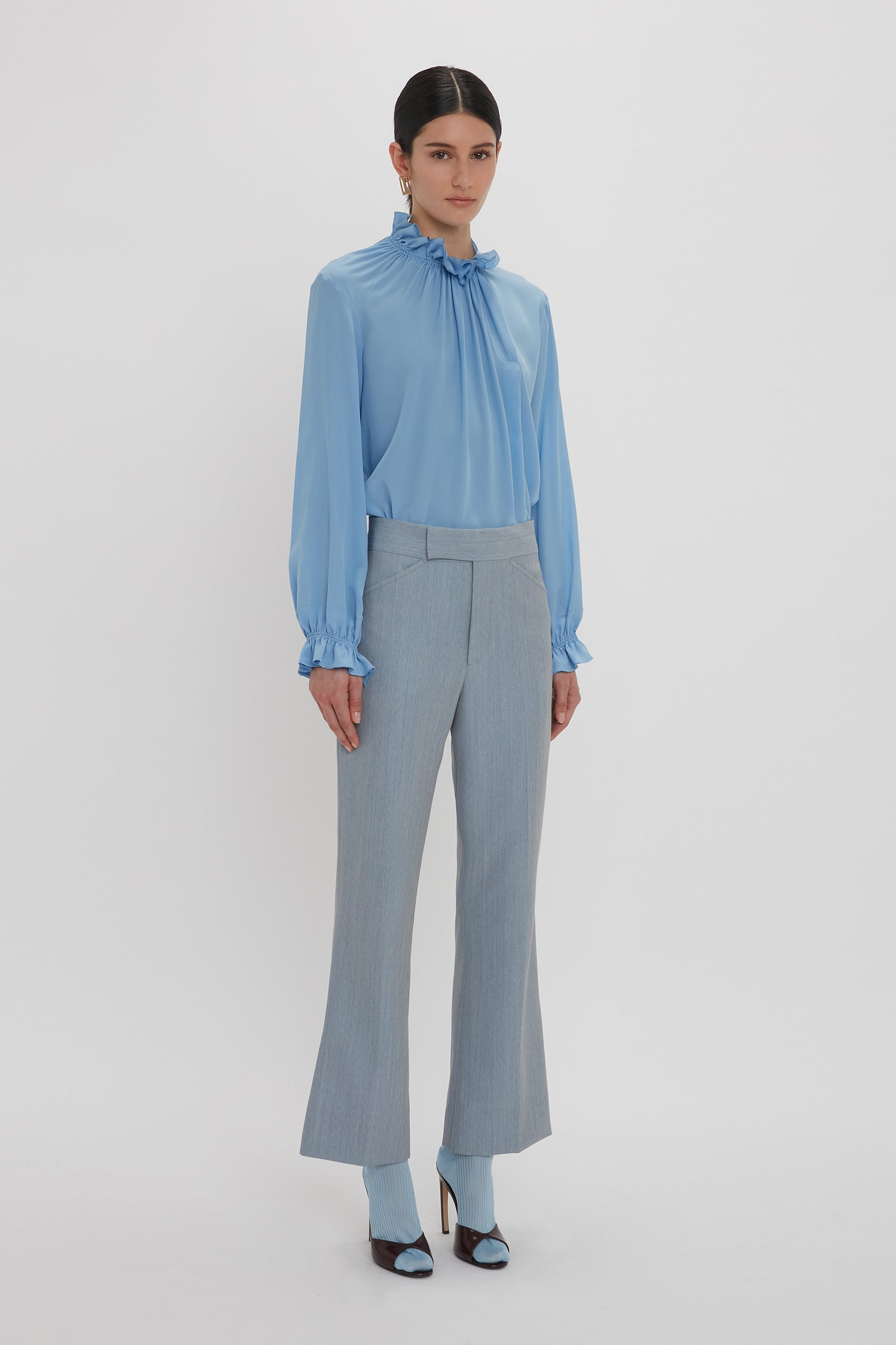 A person stands against a plain background, wearing the Victoria Beckham Exclusive Ruffle Neck Blouse In Cornflower Blue, paired with high-waisted, light gray pants and matching cornflower blue heels.