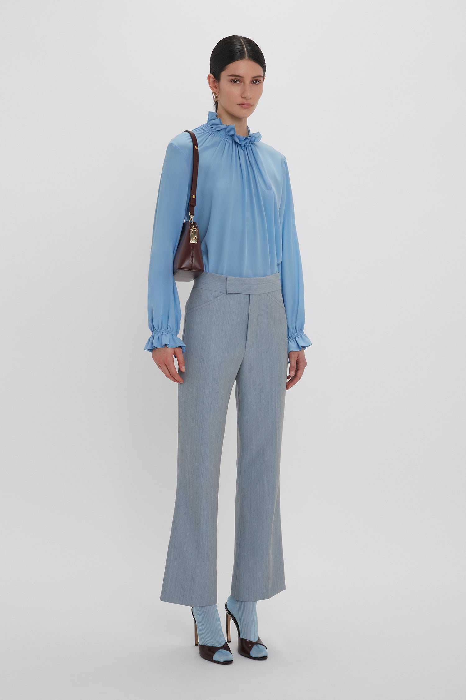 A person is standing against a white background, wearing an Exclusive Ruffle Neck Blouse In Cornflower Blue by Victoria Beckham made from crepe de chine fabric and light gray pants, holding a brown handbag over their shoulder. They have dark hair and are wearing blue high-heeled shoes.