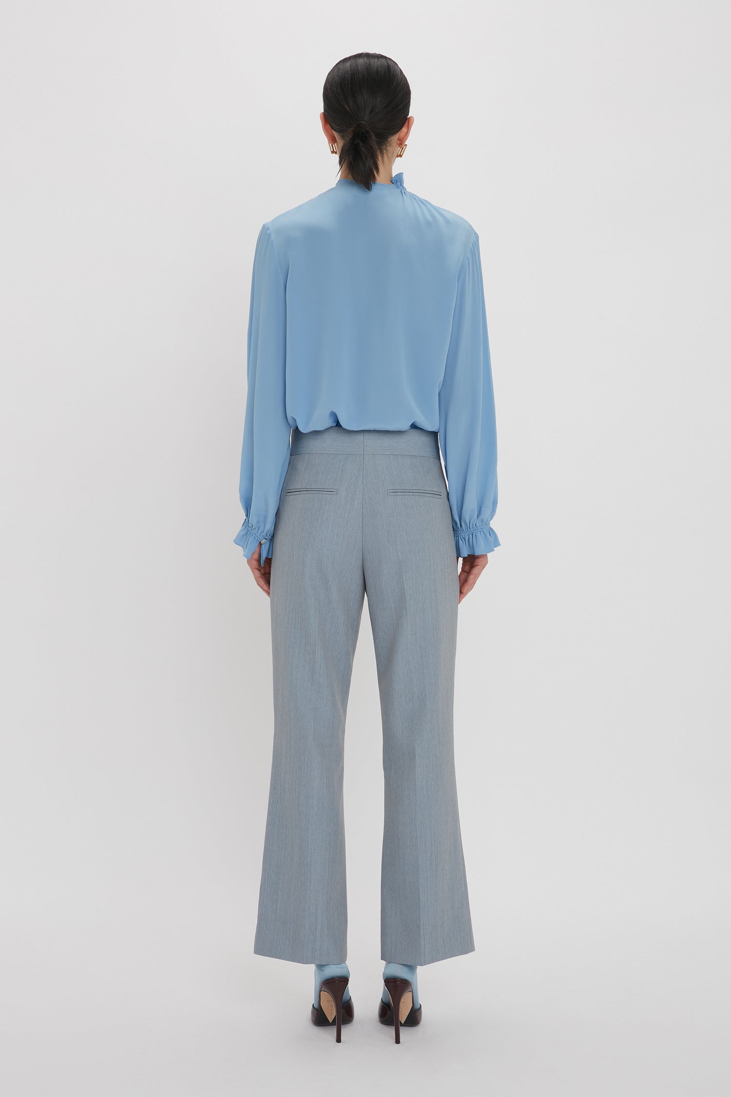 A person is standing with their back to the camera, wearing an Exclusive Ruffle Neck Blouse In Cornflower Blue from Victoria Beckham and gray pants. Their hair is tied back, and they are wearing high-heeled shoes.
