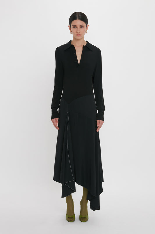 A person stands against a white background wearing a Victoria Beckham Henley Shirt Dress In Black with an asymmetric waist and olive green shoes.