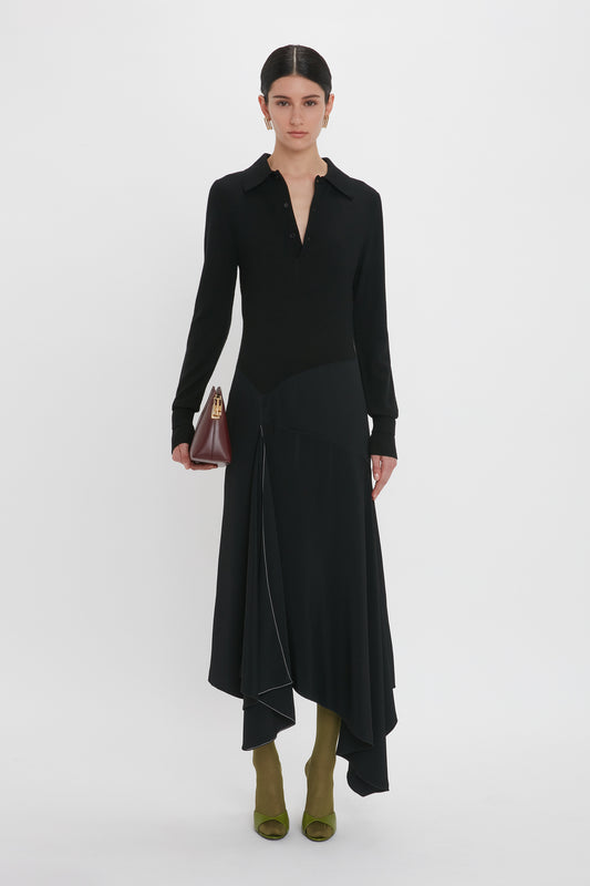 A person in a long-sleeved Henley Shirt Dress In Black by Victoria Beckham with an asymmetrical hem stands against a plain white background. They are holding a small maroon handbag and wearing olive green shoes.