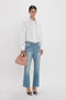 Person stands against a white background wearing a white shirt, blue jeans, and black heels, holding a W11 Mini Tote Bag In Marshmallow Leather by Victoria Beckham.