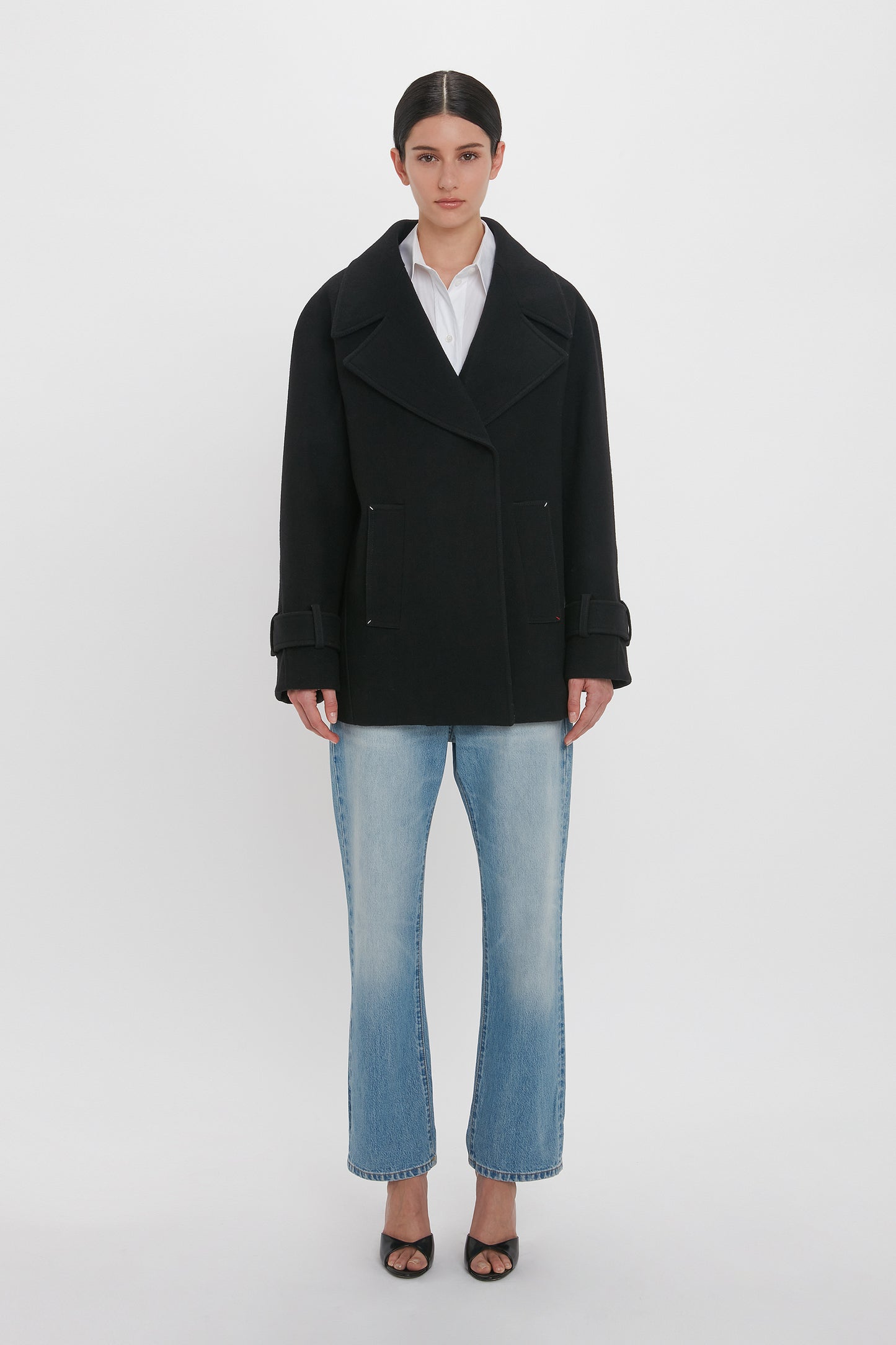 A person stands against a plain white background, wearing a black Oversized Pea Coat In Black by Victoria Beckham featuring masculine tailoring, a white shirt, light blue jeans, and black open-toe heels.