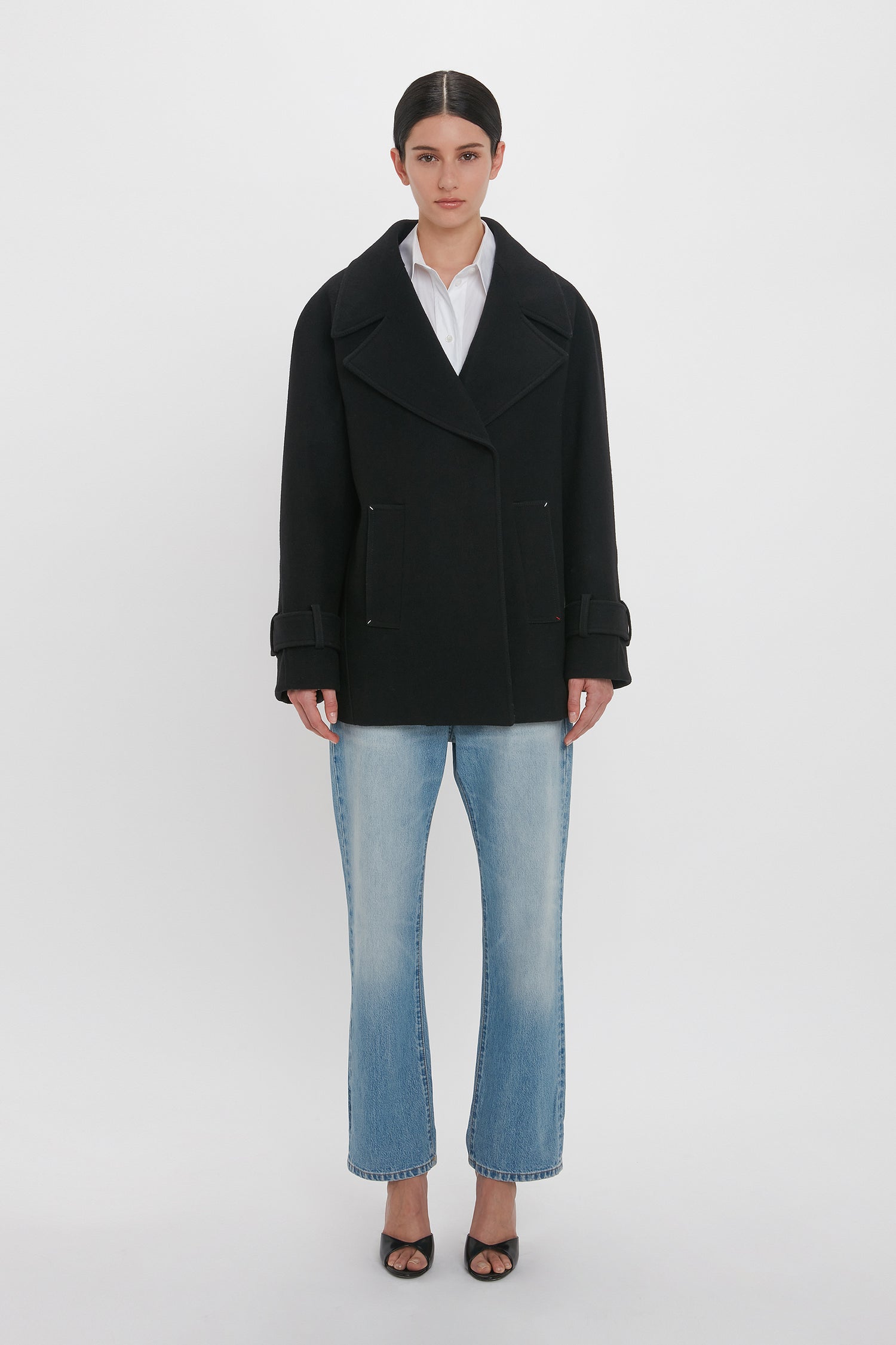 A person stands against a plain white background, wearing a black Oversized Pea Coat In Black by Victoria Beckham featuring masculine tailoring, a white shirt, light blue jeans, and black open-toe heels.