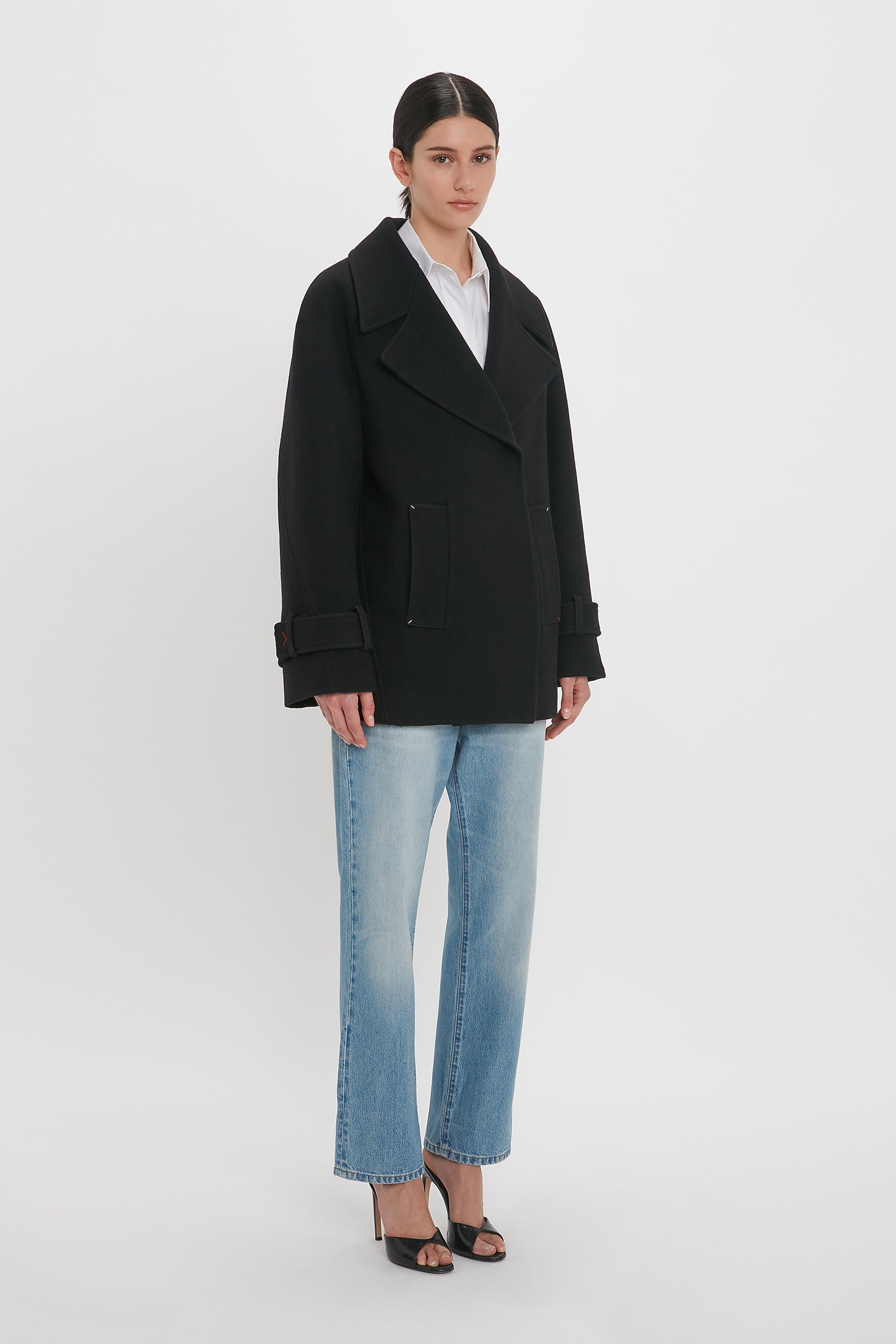 A person stands wearing a Victoria Beckham Oversized Pea Coat In Black over a white shirt, blue denim jeans, and black heeled sandals against a plain white background.