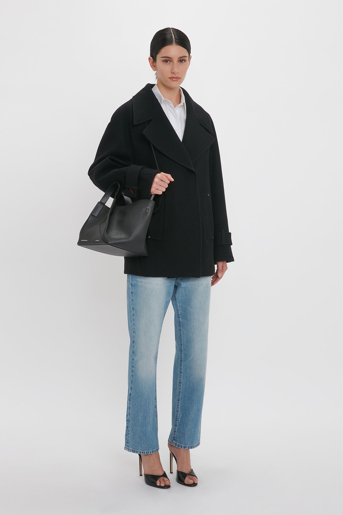 A person stands against a plain white background, wearing the Victoria Beckham Oversized Pea Coat In Black made of melton wool, light blue jeans, and black high heels, holding a black handbag.