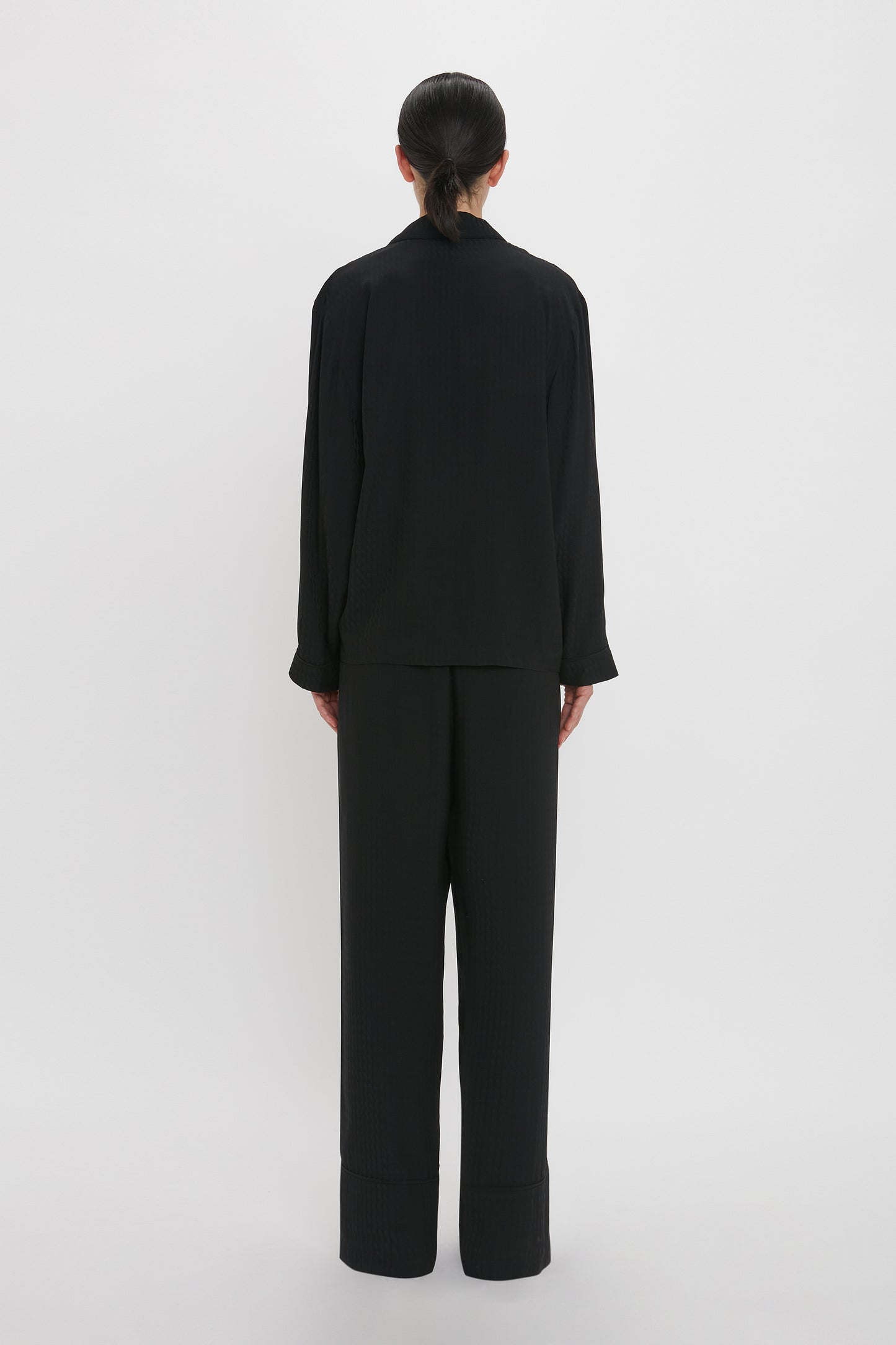 Person standing facing away, wearing a black long-sleeved shirt and black wide-leg pants that resemble the VB Monogram Pyjama Sleep Set In Black by Victoria Beckham, against a plain white background.