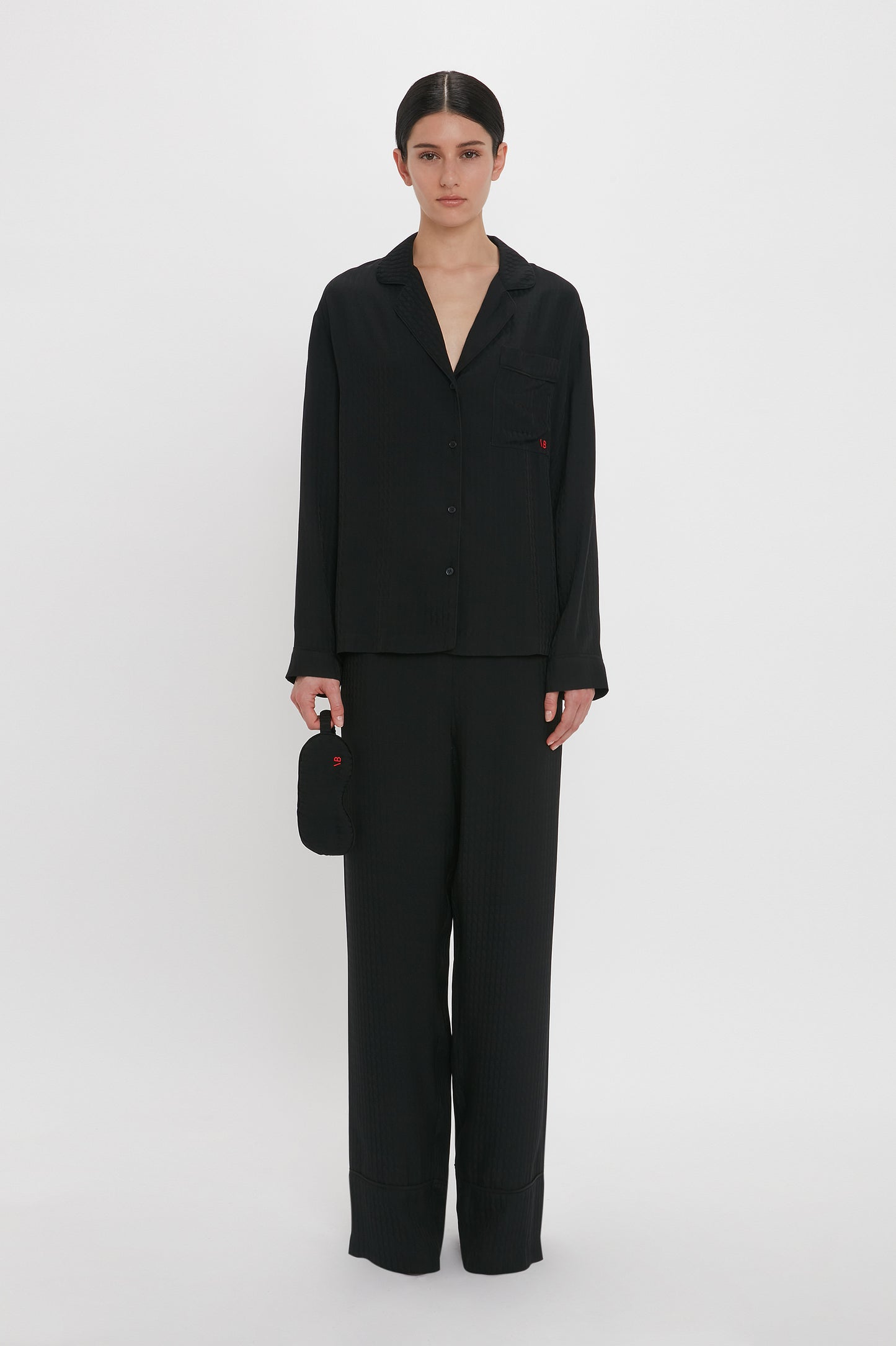 Person in black, VB Monogram Pyjama Sleep Set In Black by Victoria Beckham, standing against a plain white background, holding a small black pouch.