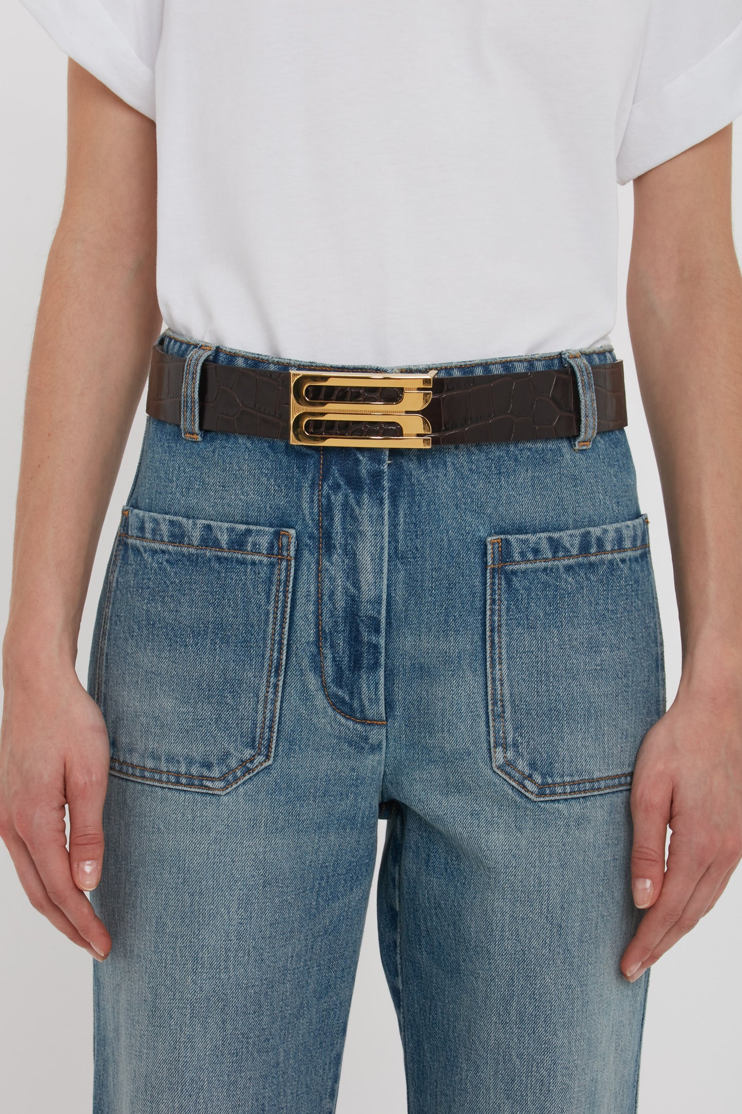 A person wearing a plain white t-shirt is shown from the waist down, donning blue jeans with large front pockets and a Victoria Beckham Jumbo Frame Belt in Espresso Croc Embossed Calf Leather featuring two luxury gold buckles.