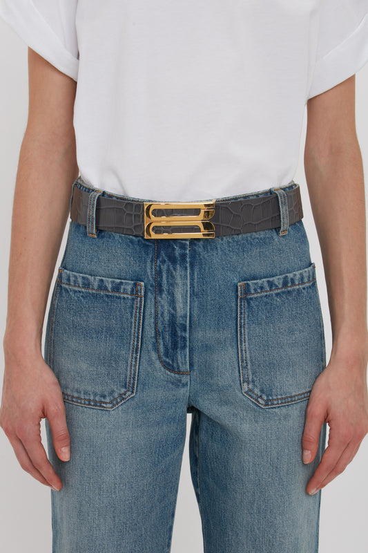 Person wearing a white shirt and blue jeans, cinched with a black belt featuring a gold buckle. The outfit is elevated by the slate grey, croc-embossed calf leather detailing on the Victoria Beckham Jumbo Frame Belt In Slate Grey Croc Embossed Calf Leather.