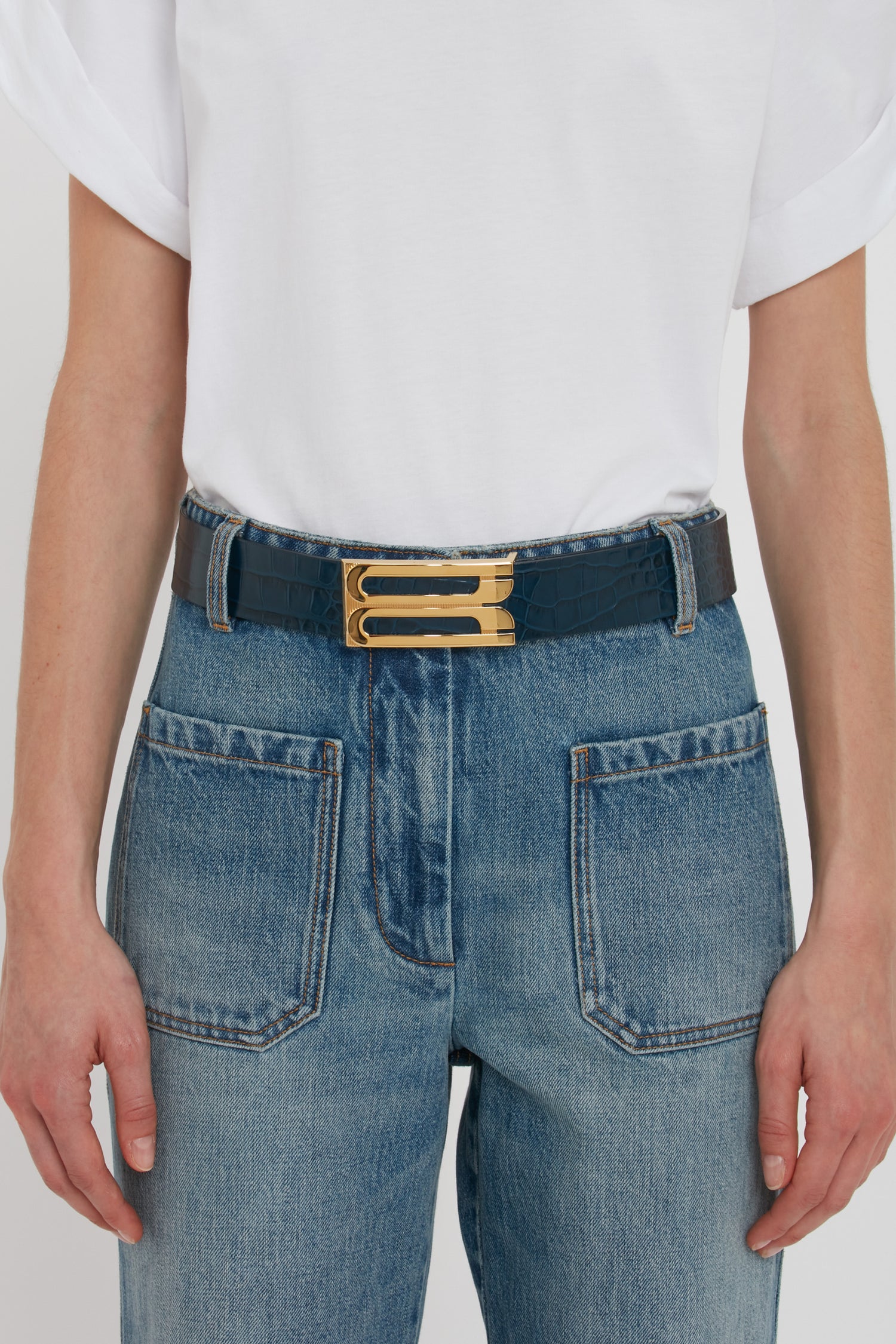 Person wearing a white t-shirt, blue jeans with large front pockets, and a black Victoria Beckham Jumbo Frame Belt In Midnight Blue Croc Embossed Calf Leather featuring luxury gold hardware.