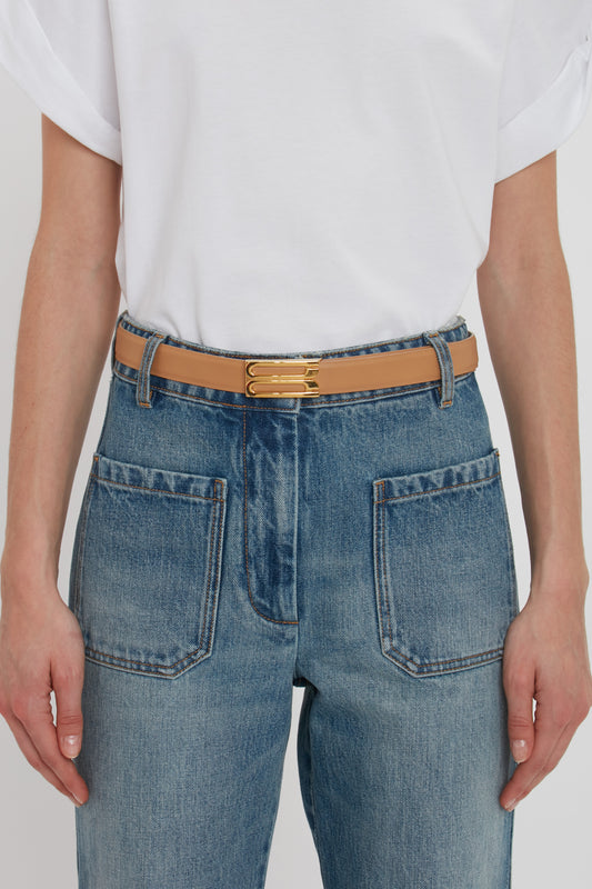 A person wearing a white t-shirt, light blue denim jeans with large pockets, and a beige Victoria Beckham Frame Belt In Camel Leather featuring luxury gold hardware.