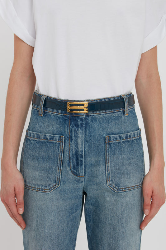 A person wearing a white t-shirt tucked into high-waisted midnight blue jeans with the Victoria Beckham Frame Belt In Midnight Blue Croc Embossed Calf Leather featuring gold hardware.