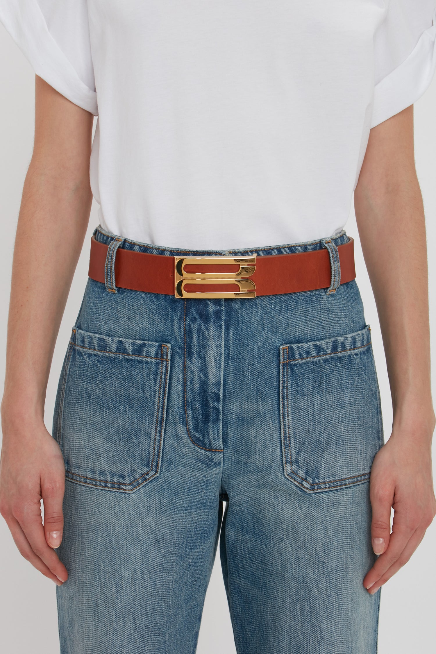 Person wearing a white short-sleeve shirt tucked into blue jeans with large front pockets, accessorized with a tan Exclusive Jumbo Frame Belt In Tan Leather featuring smooth calf leather and gold hardware by Victoria Beckham.