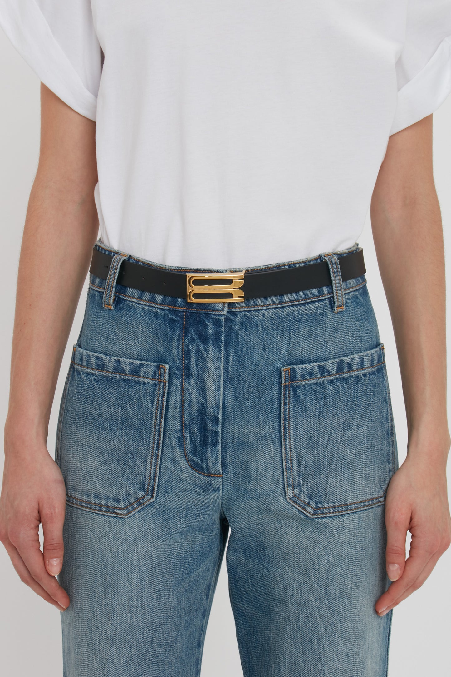 A person wearing blue jeans with oversized front pockets, a white t-shirt, and a Victoria Beckham Frame Belt In Black Leather glints in the light. The photo captures the torso and waist area, highlighting this versatile accessory that stands out as a wardrobe classic.