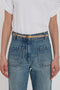 A person wearing a white t-shirt tucked into high-waisted blue jeans with large front pockets and the Victoria Beckham Exclusive Micro Frame Belt In Beige Leather with gold hardware.