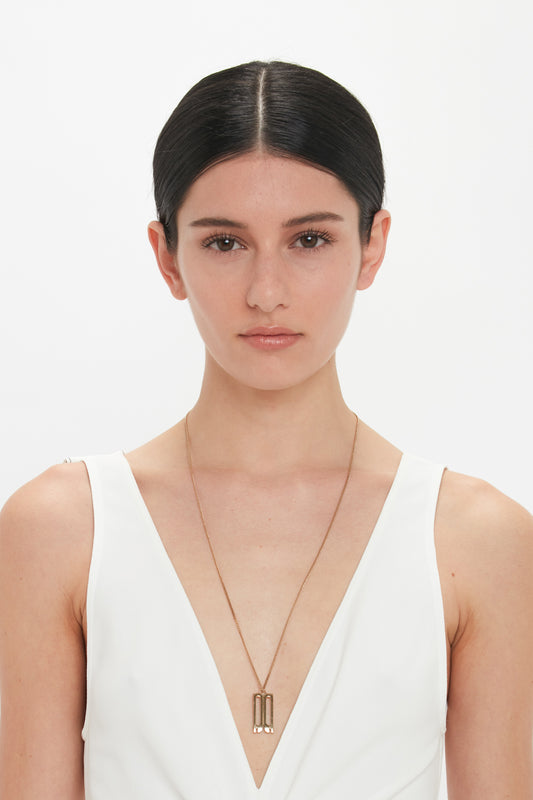 A person with straight dark hair, wearing a sleeveless white top and a long Victoria Beckham necklace with an Exclusive Frame Necklace In Gold, stands in front of a plain white background, their facial expression neutral.