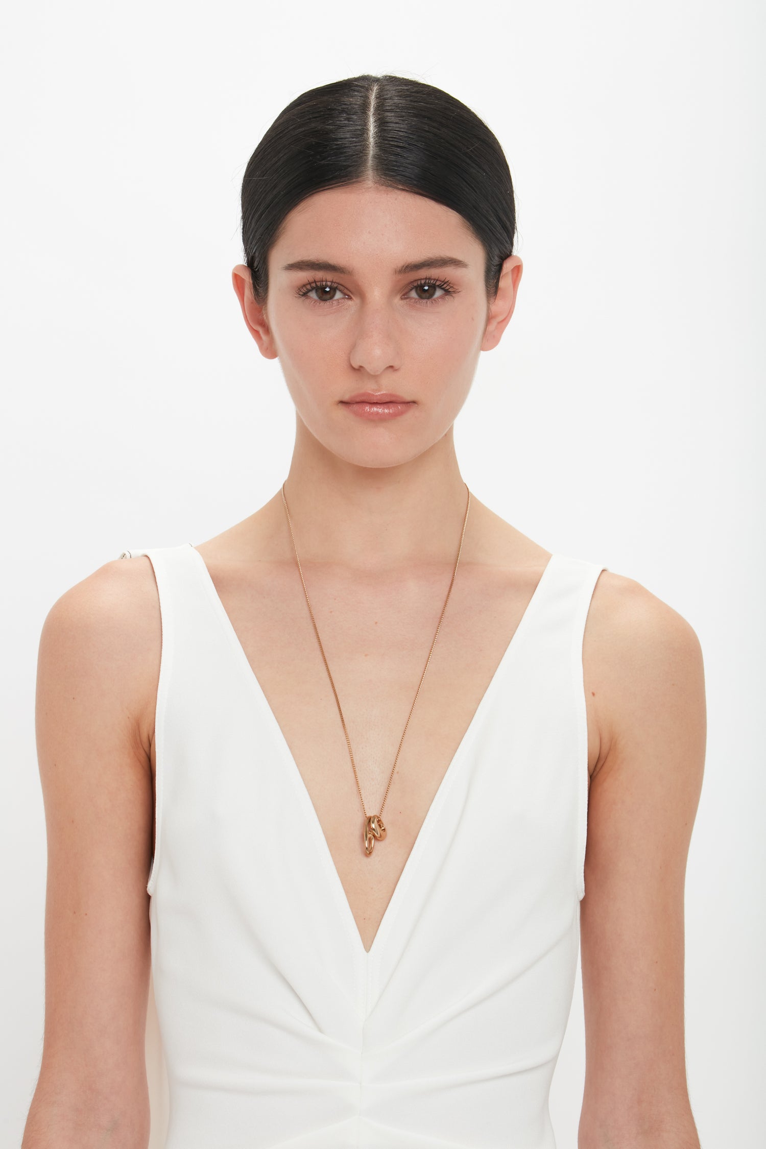 A person with dark hair in a middle part wears a white sleeveless V-neck top and an exquisitely crafted in Italy thin necklace, the Exclusive Absract Charm Necklace In Light Gold by Victoria Beckham, standing against a white background.