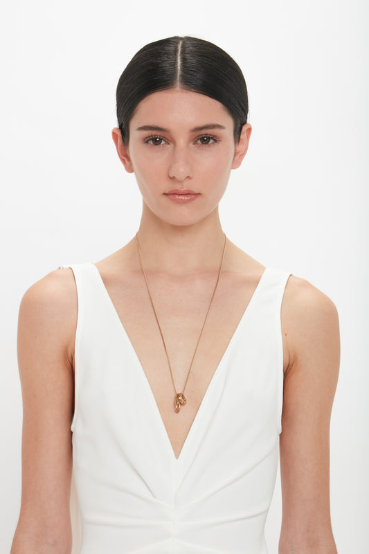 A person with dark hair in a middle part wears a white sleeveless V-neck top and an exquisitely crafted in Italy thin necklace, the Exclusive Absract Charm Necklace In Light Gold by Victoria Beckham, standing against a white background.