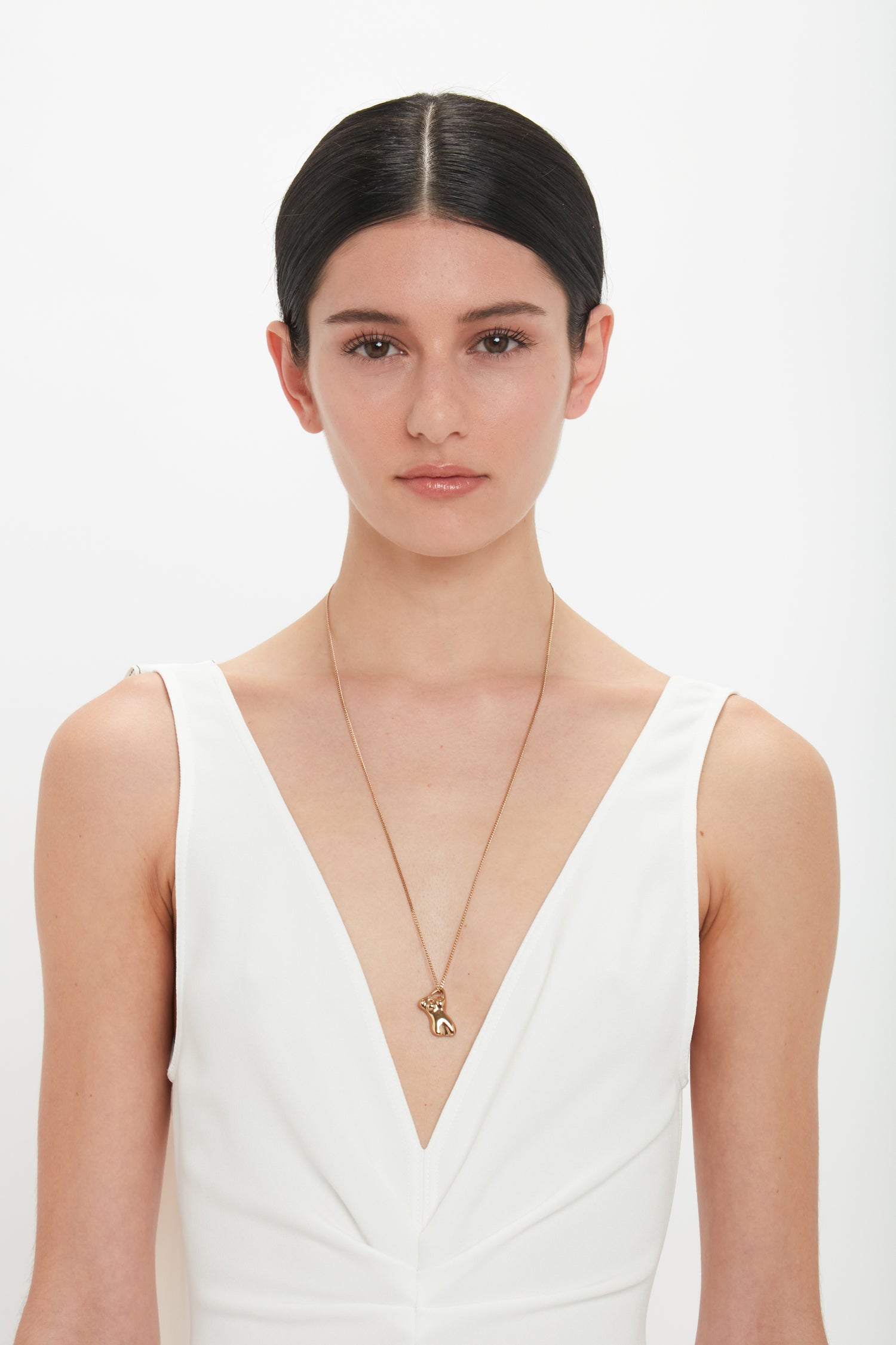 A person with straight dark hair, wearing a sleeveless white dress and an Exclusive Body Charm Necklace In Light Gold by Victoria Beckham made of 100% brass, stands against a plain white background.