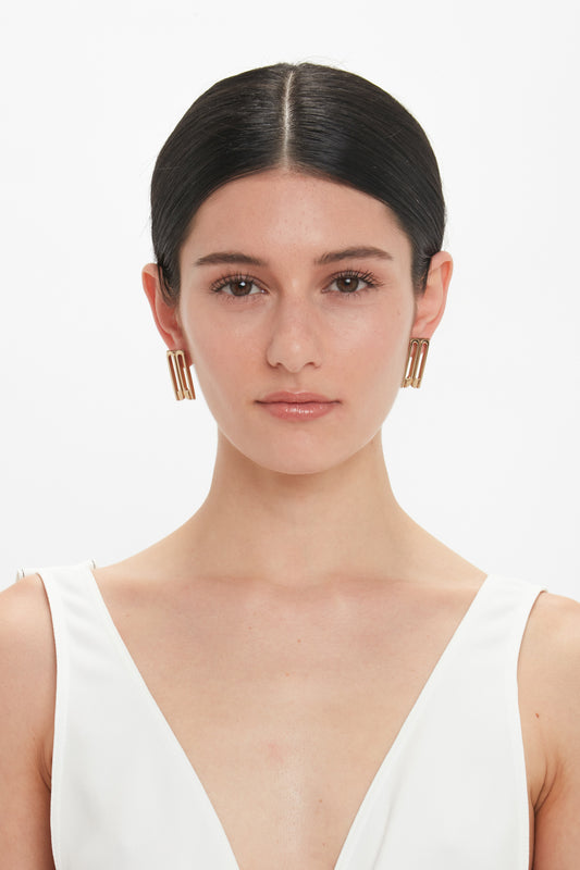 A female model with dark hair, wearing Exclusive Frame Stud Earrings In Gold by Victoria Beckham and a white v-neck top, poses against a plain white background.