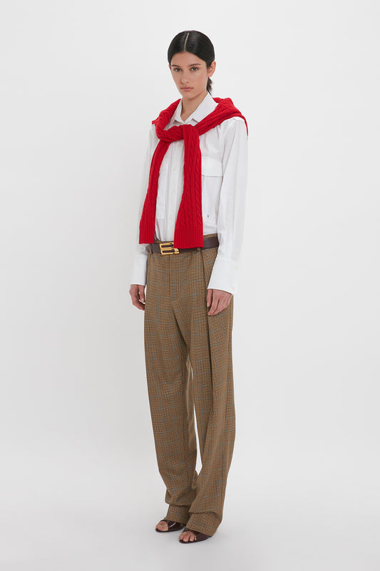 Individual wearing a white shirt, red sweater draped over shoulders, and Victoria Beckham Asymmetric Chino Trouser In Tobacco/Multi stands against a plain background.