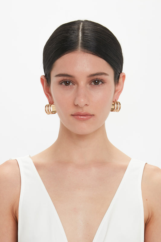 A person with straight, dark hair pulled back wearing a white top and Victoria Beckham Exclusive Frame Hoop Earrings In Gold stands against a plain white background.