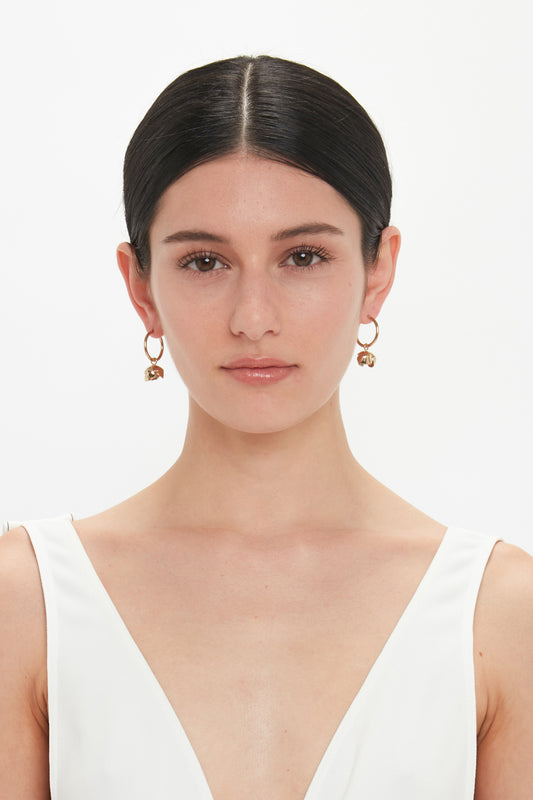 A woman with straight black hair and light makeup wears a white V-neck top and stunning Exclusive Camellia Flower Hoop Earrings In Gold by Victoria Beckham, hand-crafted in Italy, standing against a plain white background.