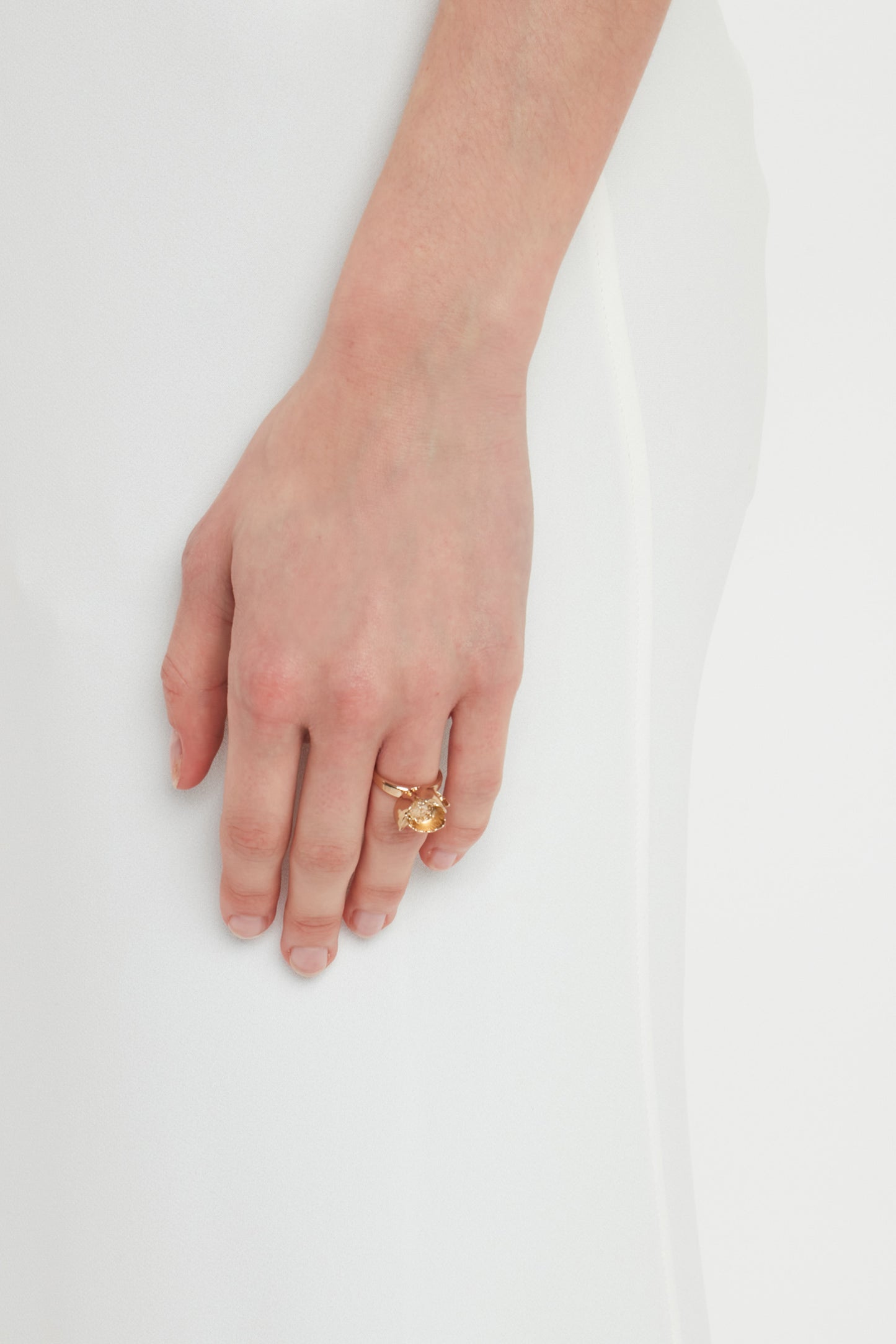 A woman’s hand wearing an Exclusive Camellia Flower Ring In Gold by Victoria Beckham, handcrafted in Italy, rests elegantly against a white fabric background.