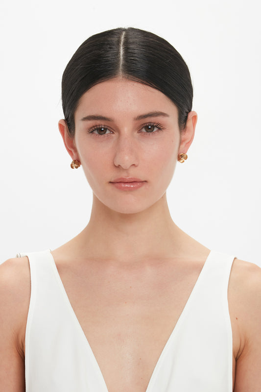 A woman with dark hair tied back, wearing a white sleeveless V-neck top and Victoria Beckham Exclusive Camellia Flower Stud Earrings In Gold, stands against a plain white background.