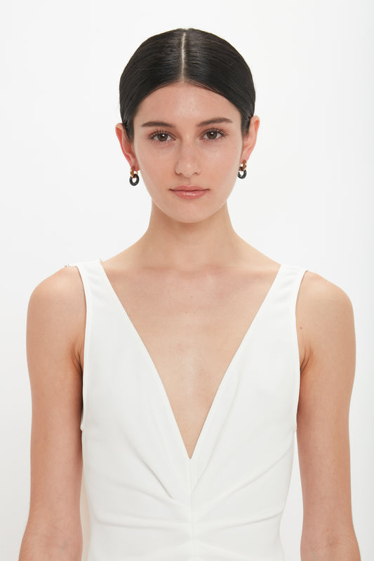 A person with dark hair, wearing a white, deep V-neck sleeveless top, stands against a plain white background, accentuated by Victoria Beckham Exclusive Resin Pendant Earrings In Light Gold-Black.