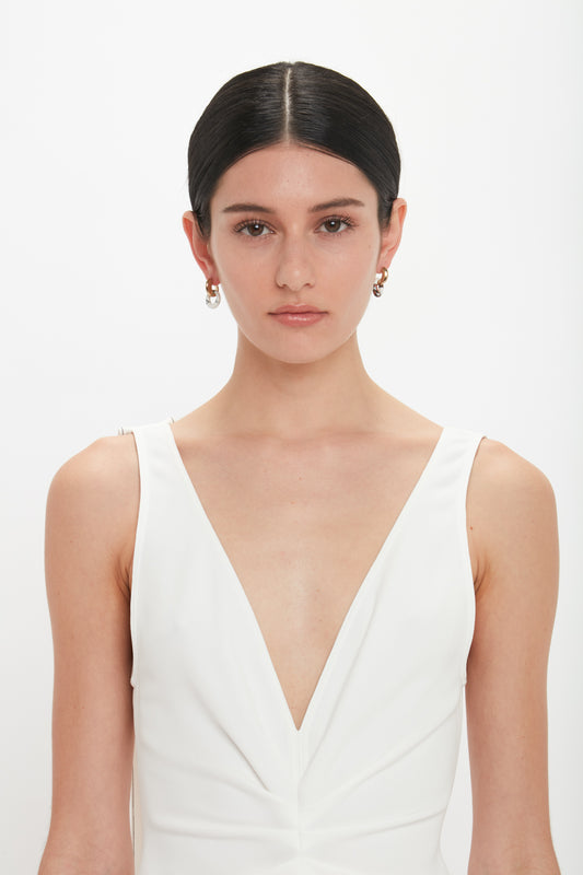 A person with dark hair, tied back, is wearing a deep v-neck white top and Victoria Beckham Exclusive Resin Pendant Earrings In Light Gold-White against a plain white background.