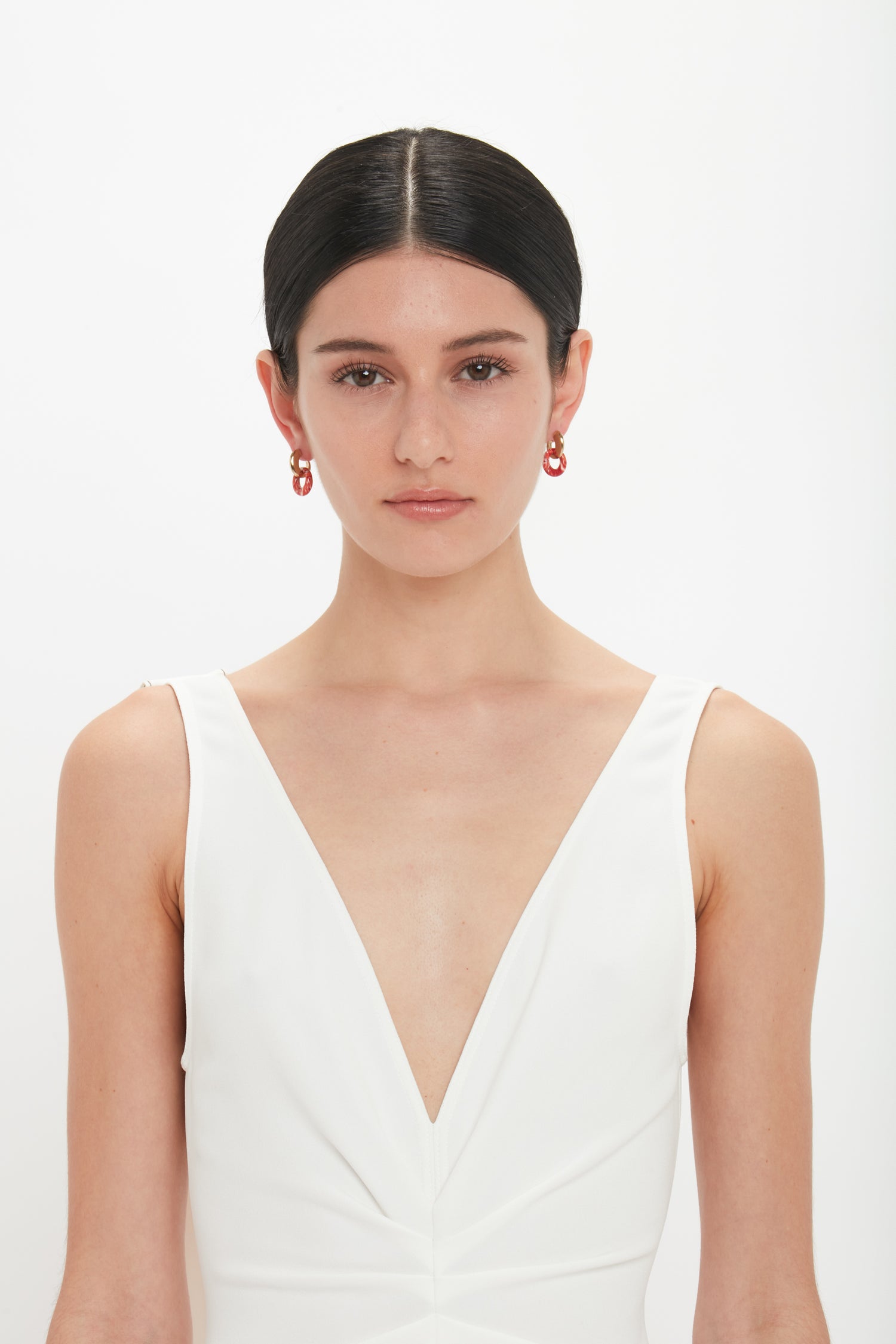 A woman with dark hair, wearing Victoria Beckham Exclusive Resin Pendant Earrings In Light Gold-Orange and a white sleeveless dress with a deep V-neckline, stands against a plain white background.