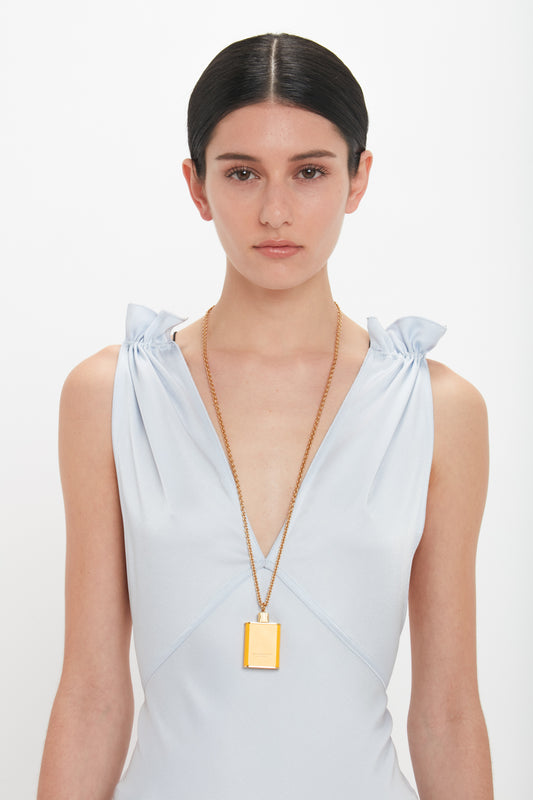 A woman with dark hair in an updo wears a light blue sleeveless dress and a long gold necklace with a rectangular pendant, reminiscent of the Perfume Bottle Necklace In San Ysidro Drive by Victoria Beckham. She is posing against a plain white background.