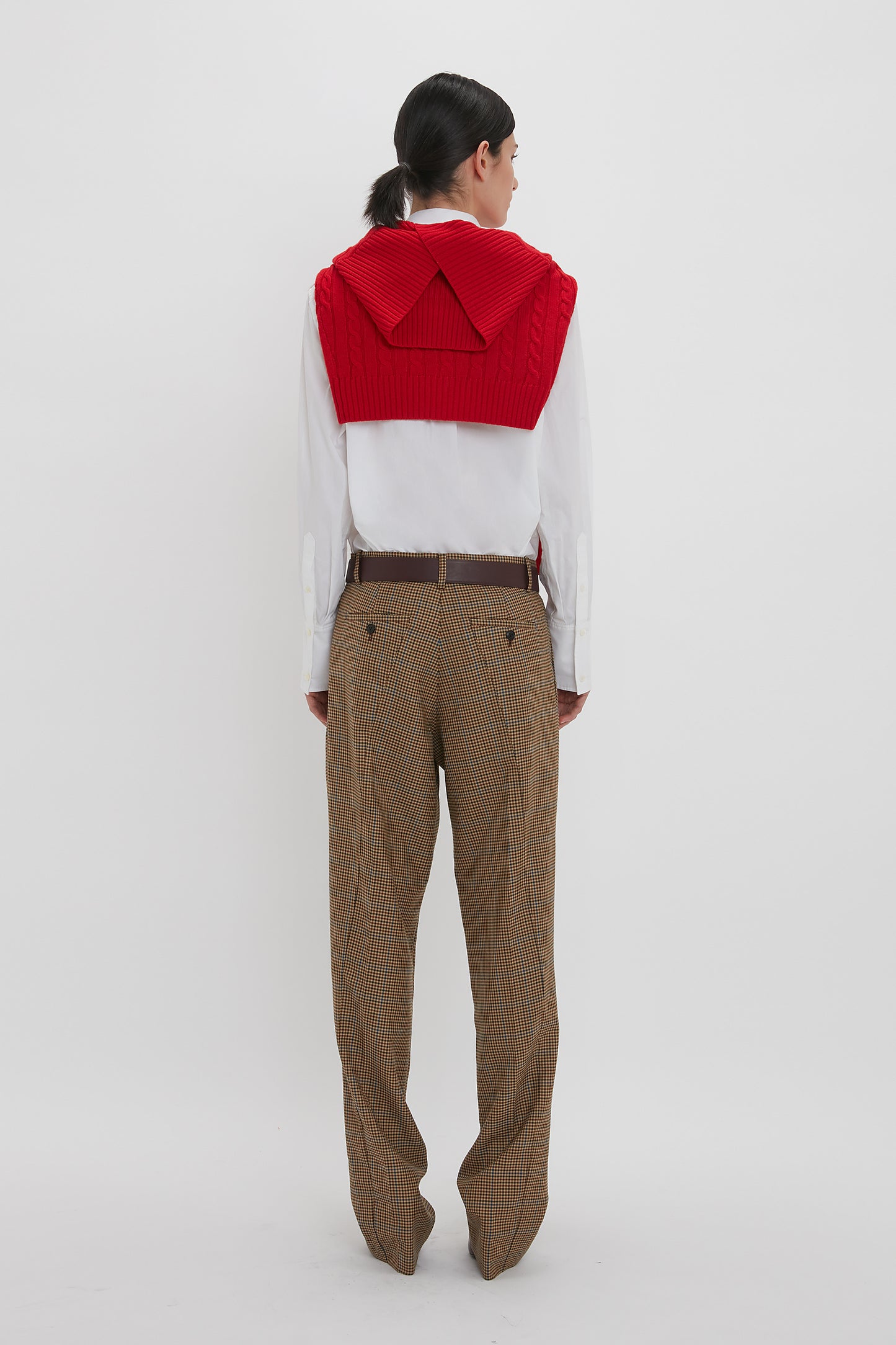 Person with long hair stands facing away, wearing a red sweater draped over a white shirt, and Victoria Beckham Asymmetric Chino Trouser In Tobacco/Multi.