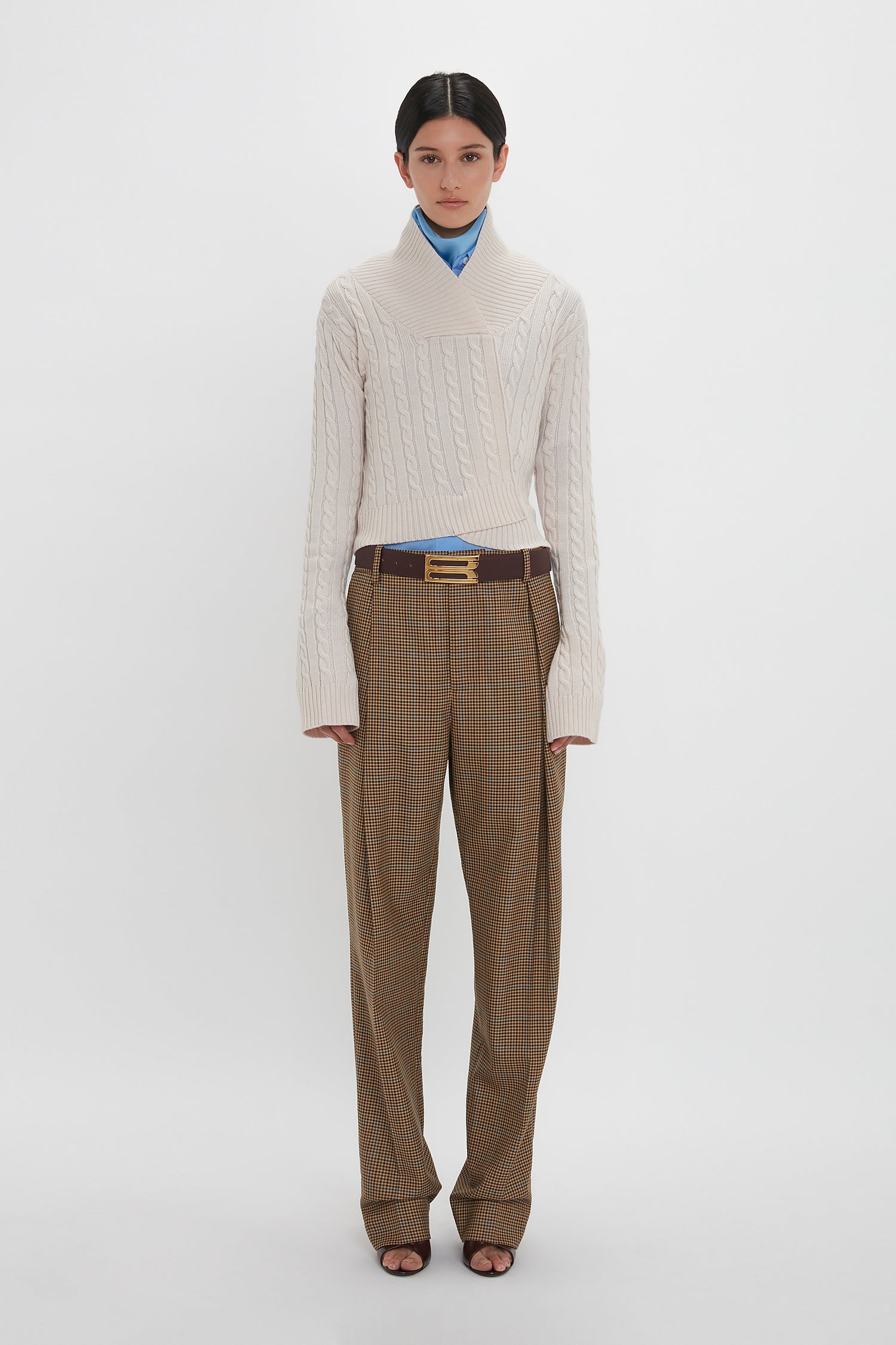 A person stands against a plain white background, adorned in luxury knitwear: the Victoria Beckham Wrap Detail Jumper In Bone and brown plaid trousers, accessorized with a brown belt and a blue scarf.