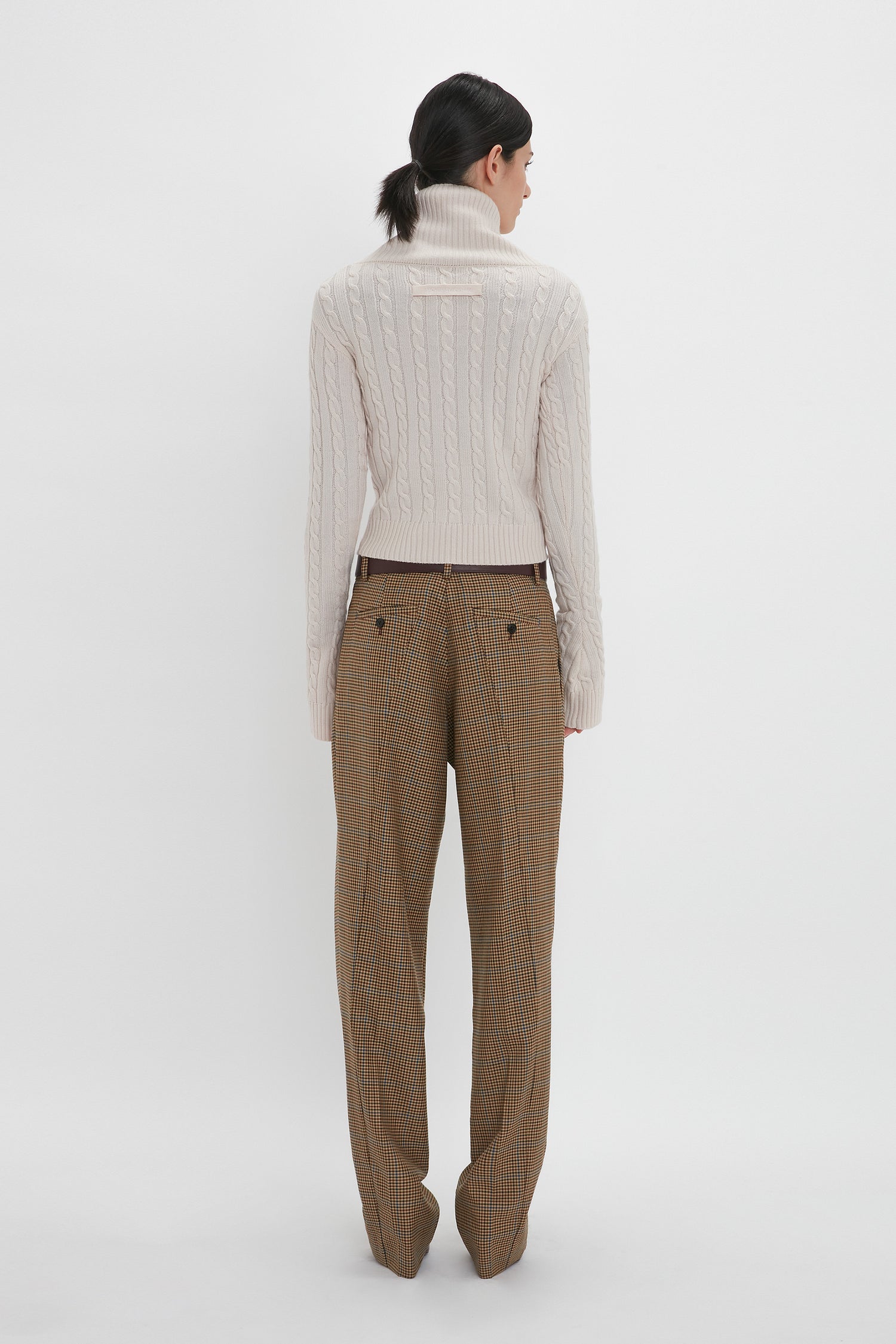  A person with dark hair, wearing a luxurious Wrap Detail Jumper In Bone and beige plaid pants from the Victoria Beckham brand, stands facing away on a white background.