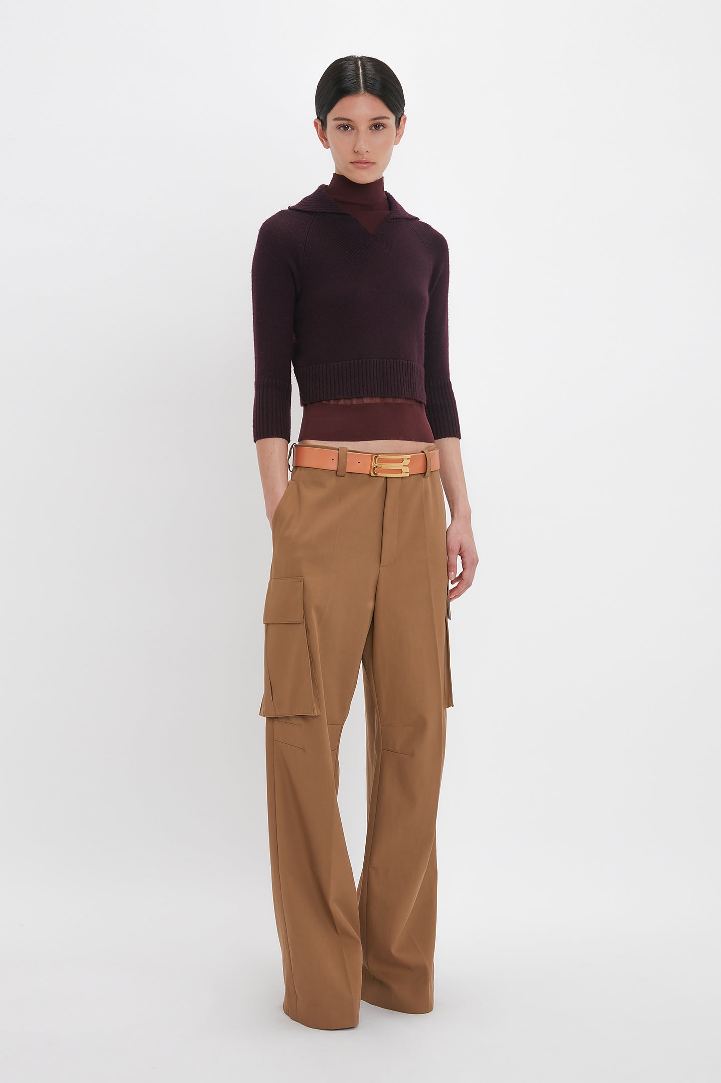 A person stands against a white background wearing the Victoria Beckham Double Layer Top In Deep Mahogany, high-waisted tan cargo pants with large pockets, and a brown belt with a gold buckle.