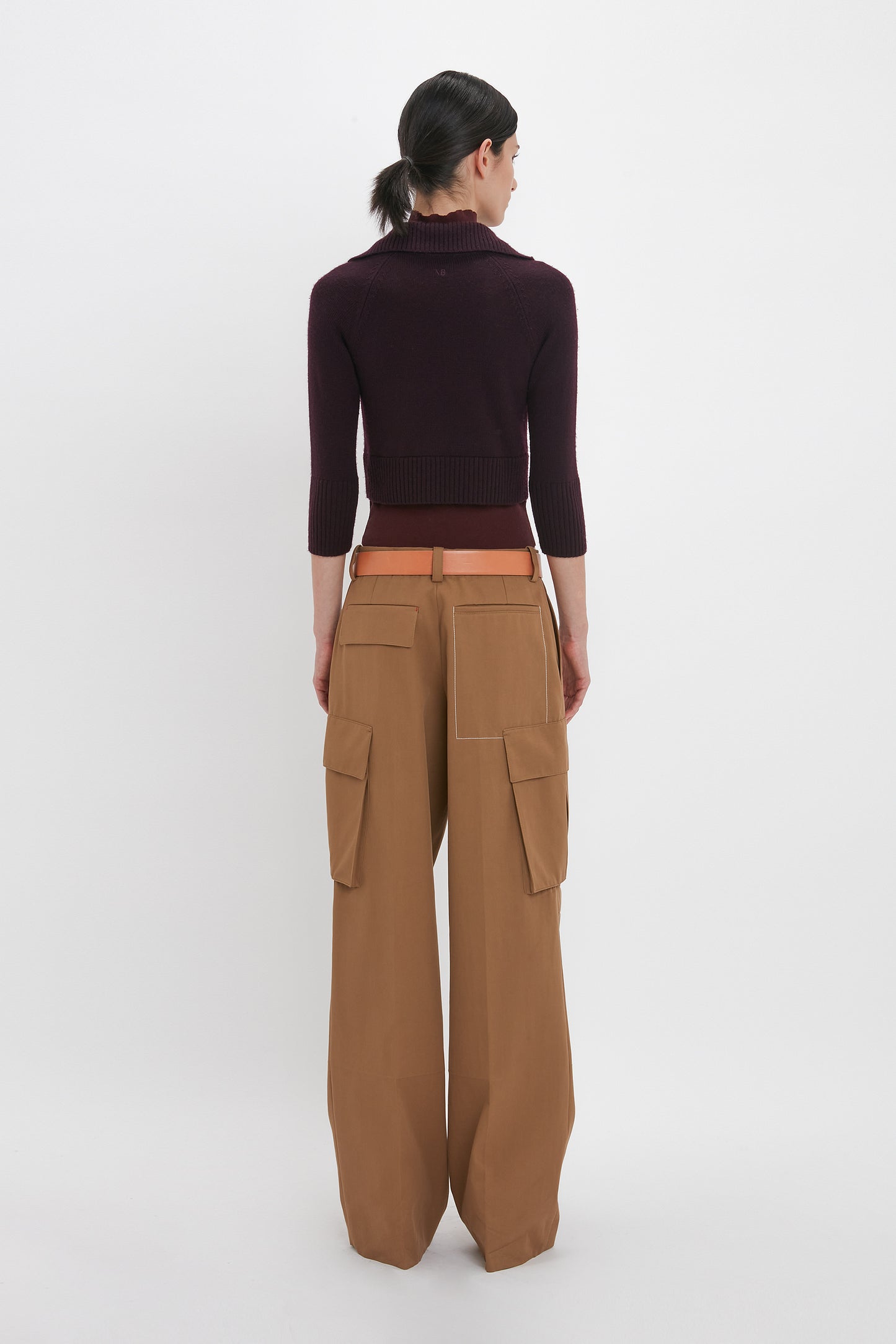 A person with dark hair in a low ponytail is standing facing away, wearing a fitted Double Layer Top In Deep Mahogany by Victoria Beckham and tan wide-leg cargo pants with pockets.