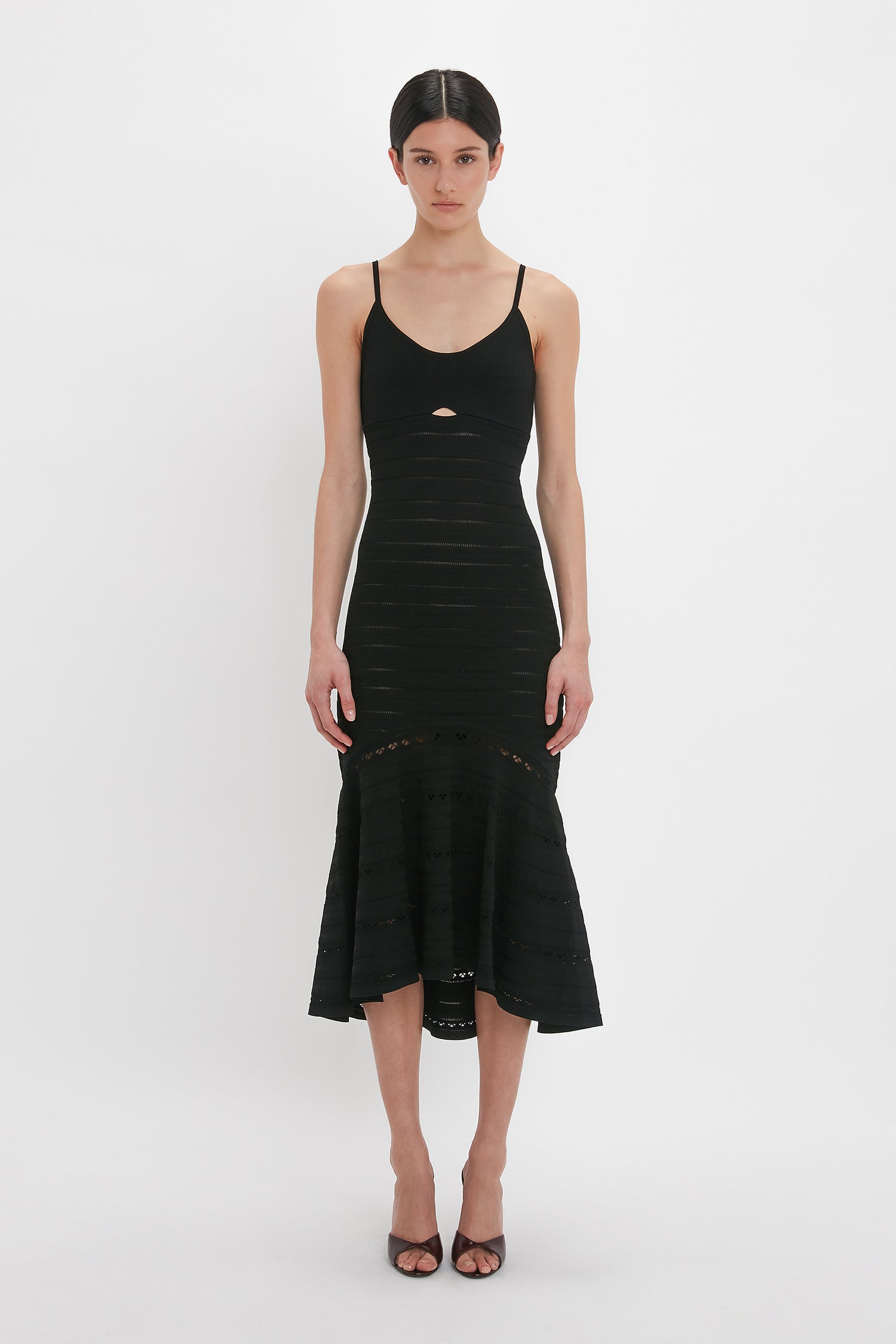 A person models the Victoria Beckham Cut-Out Detail Cami Dress In Black, a fitted black, body-sculpting dress with spaghetti straps and mid-length hem, featuring a small cutout detail below the chest. They are standing against a plain white background.