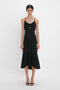 A person models the Victoria Beckham Cut-Out Detail Cami Dress In Black, a fitted black, body-sculpting dress with spaghetti straps and mid-length hem, featuring a small cutout detail below the chest. They are standing against a plain white background.