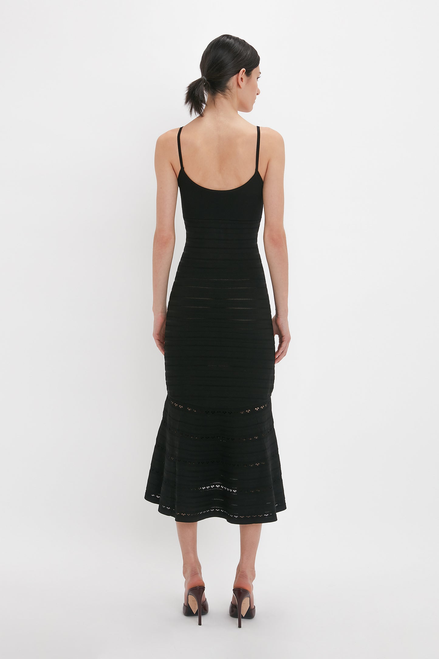 A woman with dark hair in a ponytail is seen from the back wearing the Cut-Out Detail Cami Dress In Black by Victoria Beckham with high heels, standing against a plain white background. The mid-weight stretch knit fabric and subtle cut-out detail enhance its elegance.