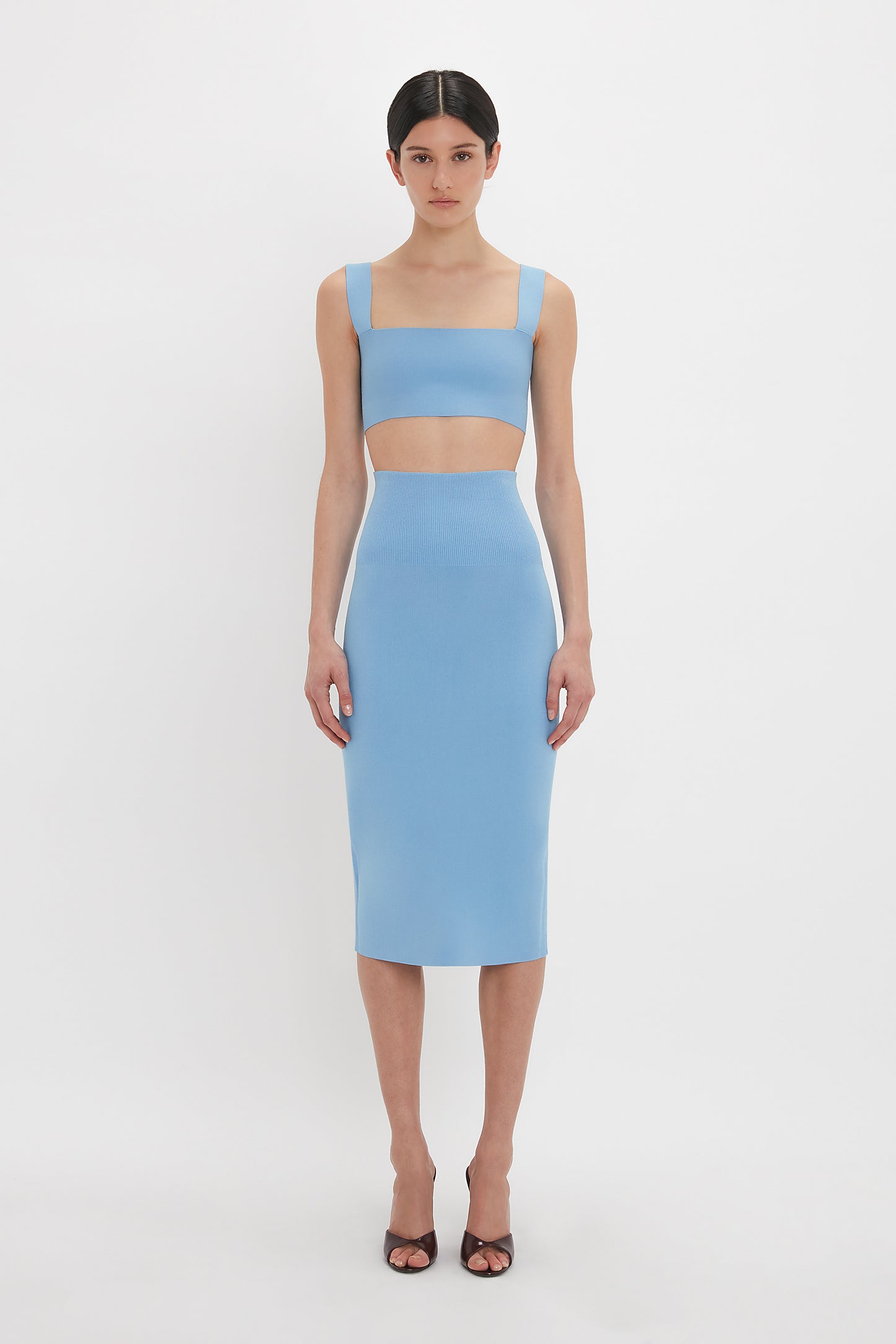 A woman stands against a white background wearing a blue, sleeveless, two-piece outfit consisting of the Strap Bandeau Top In Marina by Victoria Beckham and a fitted midi skirt, paired with black open-toe heels.