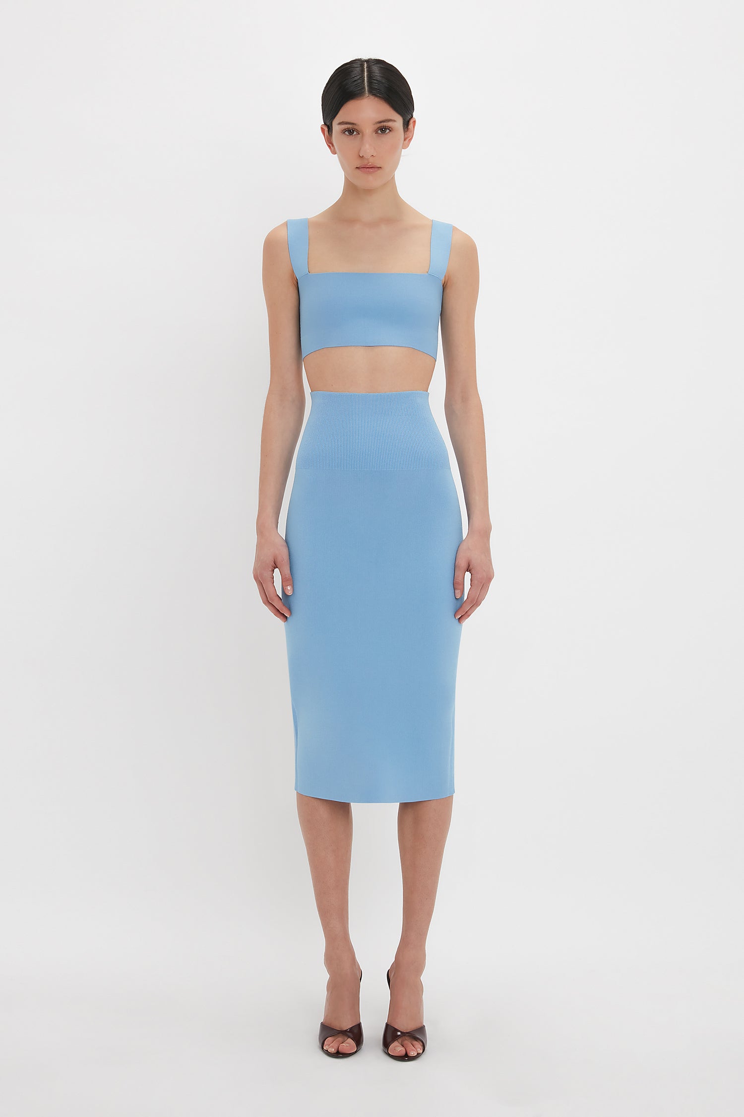 A woman stands against a white background wearing a blue, sleeveless, two-piece outfit consisting of the Strap Bandeau Top In Marina by Victoria Beckham and a fitted midi skirt, paired with black open-toe heels.