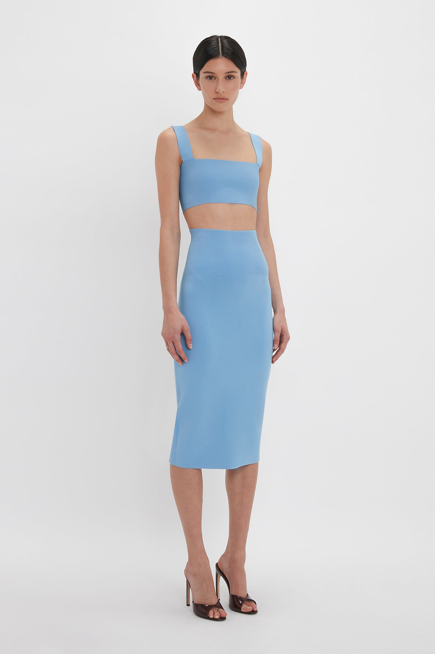 A person stands in front of a white background wearing a blue fitted midi skirt and a matching Victoria Beckham Strap Bandeau Top In Marina, paired with black high-heeled sandals.