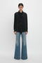 A person stands against a plain background, wearing the Victoria Beckham Asymmetric Ruffle Blouse In Black with high-waisted flared jeans. They have their hair tied back and are looking straight ahead, embodying contemporary timeless fashion.